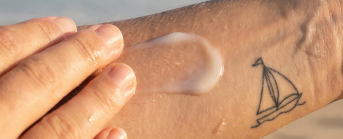 Persona che applica una crema solare su un tatuaggio al braccio in spiaggia, per proteggere la pelle dal sole e preservare il colore.